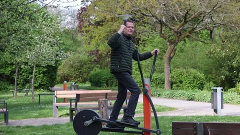 Middle-aged man exercising on outdoor elliptical machine in a park Stock Footage 334608773
