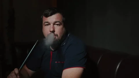 A middle-aged man exhales clouds of smoke into the camera. Dark interior of a Stock Footage 253382118