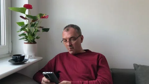 A middle-aged man with glasses over a cup of coffee sits near a window and reads Stock Footage 231748298