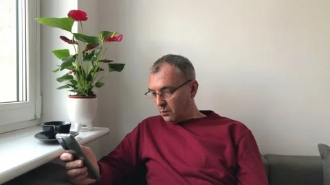 A middle-aged man with glasses over a cup of coffee sits near a window and reads Stock Footage 231748341