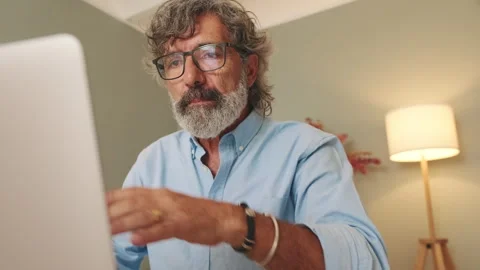 Middle-aged man with glasses sitting in the living room working on laptop Stock Footage 253928618