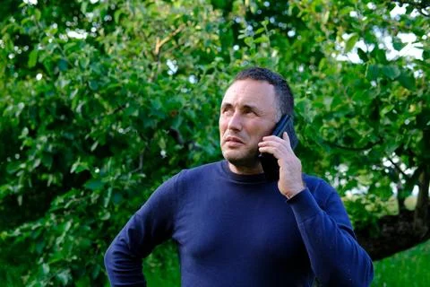 Middle aged man having serious phone conversation outdoors near green trees Stock Photos