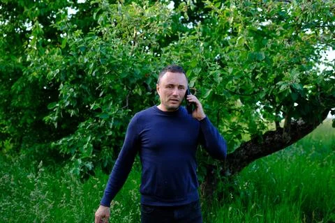 Middle aged man having serious phone conversation outdoors near green trees Stock Photos