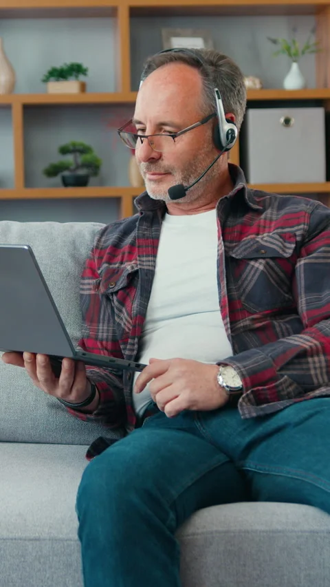 Middle aged man in headphones, talking video call, sitting on the sofa at home w Stock Footage 334439106
