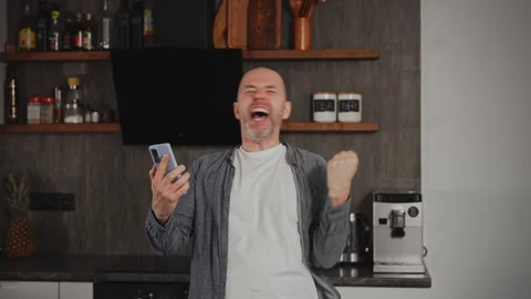 A middle-aged man is in his kitchen when he suddenly receives great news on his Stock Footage 295360174