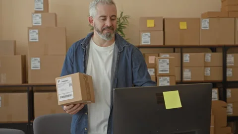 Middle aged man holds parcel and taps computer while smiling in a building;.. Stock Footage 337437025