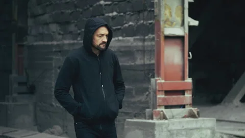 Middle-aged man in hoodie walking through industrial area, suspicious activity Stock Footage 337642003