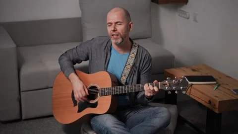 A middle-aged man in jeans and a shirt sits on the floor, playing an acoustic Stock Footage 295339963