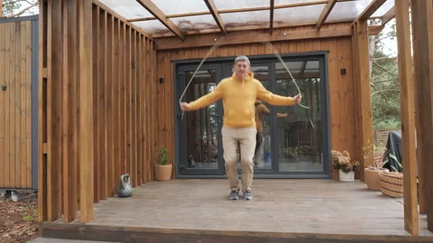Middle aged man jumping rope on wooden terrace near modern cabin Stock Footage 324704422