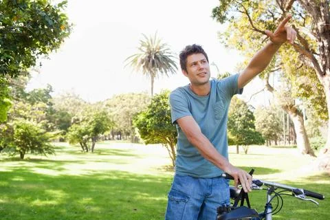 Middle-aged man leaning on bicycle and pointing toward palm tree in sunlit Photos