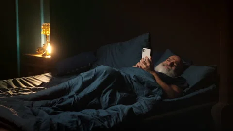 Middle-aged man lies in bed scrolling on phone, struggling with insomnia Stock Footage 346714422