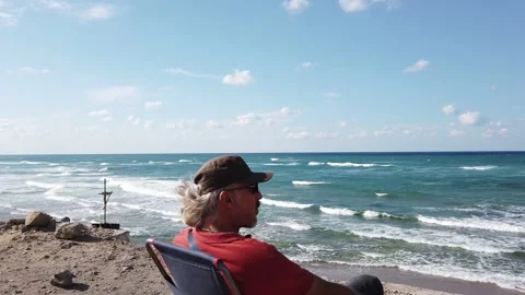 Middle-aged man with long gray hair, in dark glasses, sits resting on seashore Stock Footage 139440509
