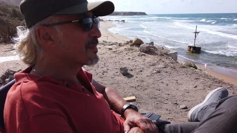 Middle-aged man with long gray hair, in dark glasses, sits resting on seashore Stock Footage 139440522