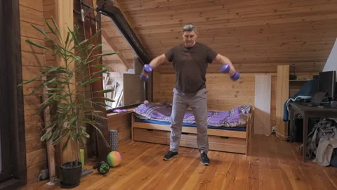 Middle aged man performing dumbbell lateral raises inside wooden attic room Stock Footage 324704386