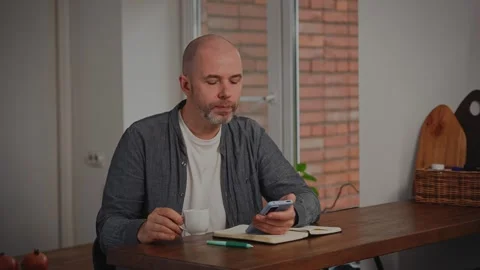 A middle-aged man relaxes in his kitchen, enjoying a cup of espresso coffee. He Stock Footage 297050165