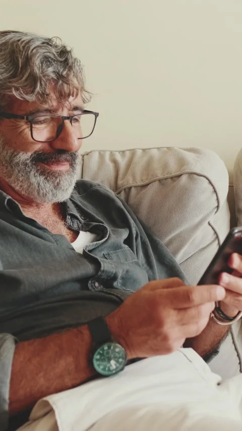 Middle-aged man relaxing on his couch while using his smartphone to text 스톡 동영상 229507833