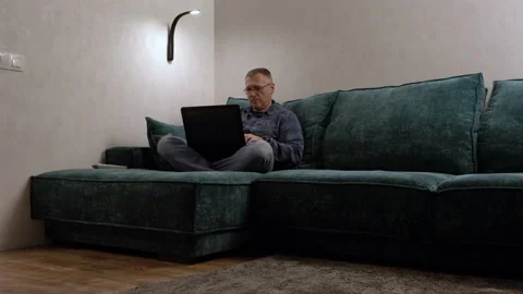 Middle-aged man relaxing on a large sofa with his laptop Stock Footage 158932947