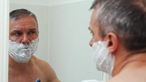 Middle aged man shaving his face with manual razor in front of mirror at home. Stock Footage 343890100