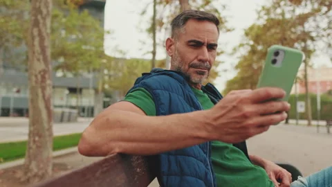 Middle-aged man sits on bench in the park uses mobile phone Stockbeeldmateriaal 242042607
