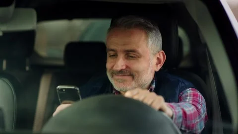 A middle-aged man sits in a car and looks at the smartphone screen. A gray Stock Footage 242609634