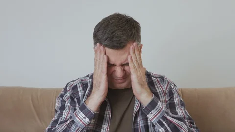A middle aged man sits on a couch and holds his hands over his head, suffering Stock Footage 104800076