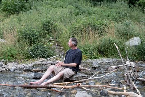 Middle aged man sits on a rock in a cool stream in the mountains contemplatin Stock Photos