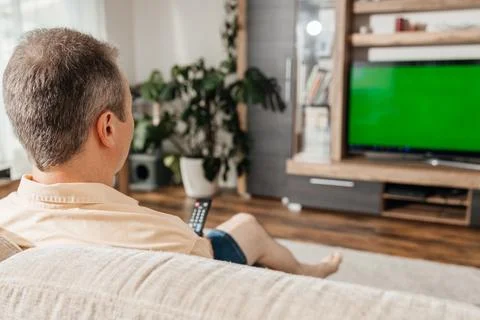 A middle-aged man sits on a sofa in the living room and watches TV with a remote Stock Photos