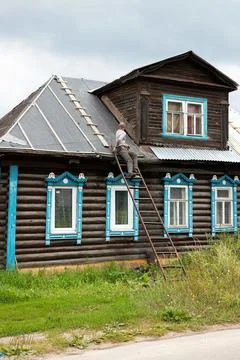 Middle-aged man sitting back on metal ladder repairing roof of old wooden brown Foto stock