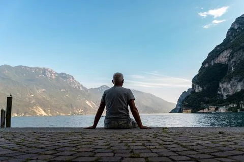 Middle-aged man sitting on the dock of a scenic harbor facing a European lake Foto stock