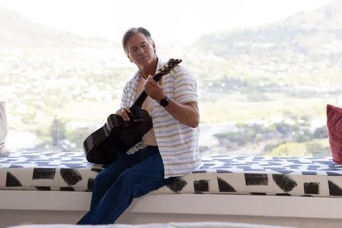 Middle-aged man sitting on patterned bench by floor-to-ceiling window, playing Foto stock