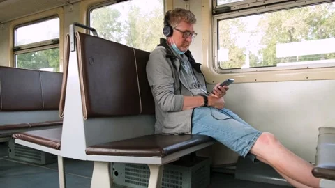 Middle-aged man is sitting on public transport with headphones and using phone. Video stock 159541629