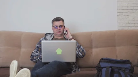 A middle-aged man sitting on a sofa is using a laptop and talking on the phone Stock Footage 104593896