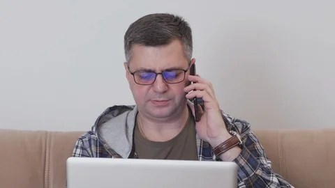A middle-aged man sitting on a sofa is using a laptop and talking on the phone. Stock Footage 104598597