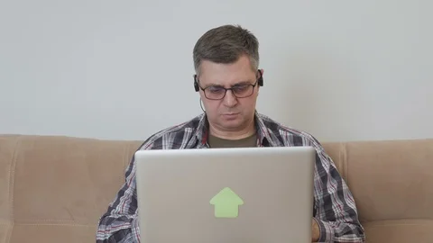 A middle-aged man sitting on a sofa with headphones in his ears enjoys a laptop Stock Footage 104786393