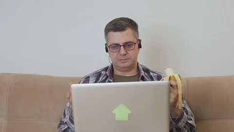 A middle-aged man sitting on a sofa with a laptop eats a banana and communicates Video stock 104843314
