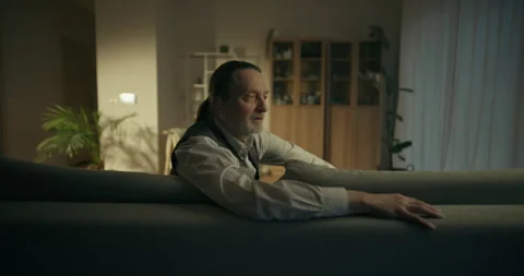 Middle-aged man sitting on a sofa in a softly lit living room 스톡 동영상 336278693