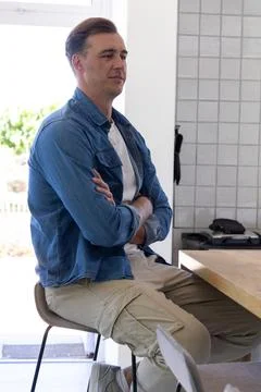 Middle-aged man sitting on stool at wooden table in bright kitchen, wearing Stock Photos