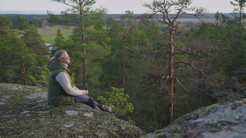 A middle-aged man is sitting on top of a cliff enjoying the sunset Stock Footage 303781790