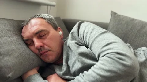 A middle-aged man sleeps on the couch wearing wireless headphones. Tired and a Stock Footage 236394277