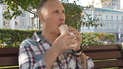Middle-aged man in square on bench eats large sandwich 스톡 동영상 120508640