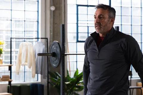 Middle-aged man standing in loft-gym wearing zip-up top by windows, barbell Stock Photos