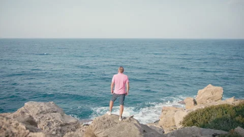 Middle Aged Man Standing on Rocky Shore Facing the Sea Stock Footage 329951263