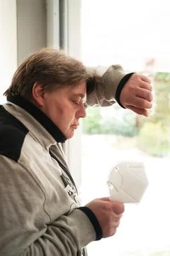 A middle aged man stands at the window looking at an FFP 2 mask. He is worrie Stock Photos