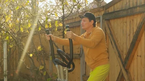 A middle-aged man uses an elliptical trainer to exercise in his yard Stock Footage 328195823