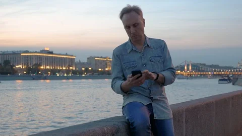 Middle aged man uses mobile phone on embankment in dusk Video stock 109607631