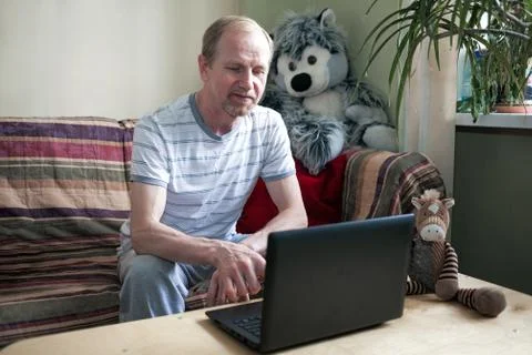 Middle aged man using a laptop computer sits at home at the table, on sofa Stock Photos