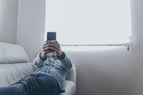 Middle aged man is using a smart phone and smiling while sitting on sofa at h Stock Photos