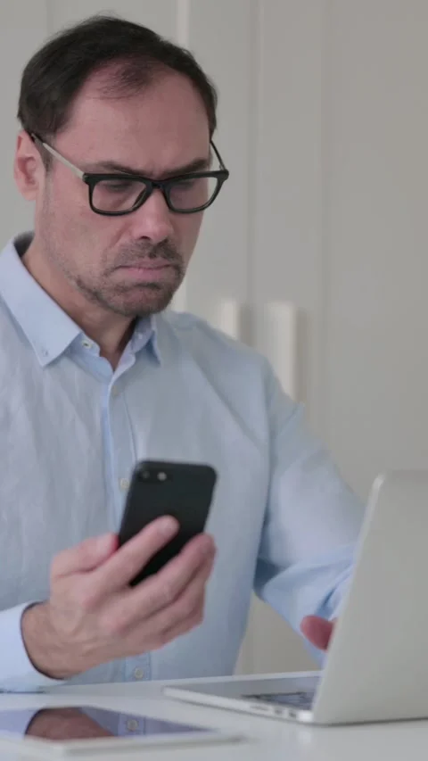 Middle Aged Man using Smartphone while working on Laptop, Vertical Video 스톡 동영상 339584760