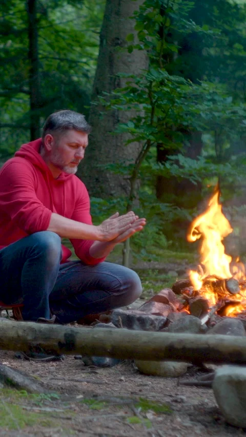 Middle aged man warming hands near campfire in forest outdoors Vidéo 334771870