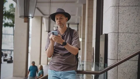 A middle-aged man wearing a hat and backpack stands in the city, using his Stock Footage 307276109